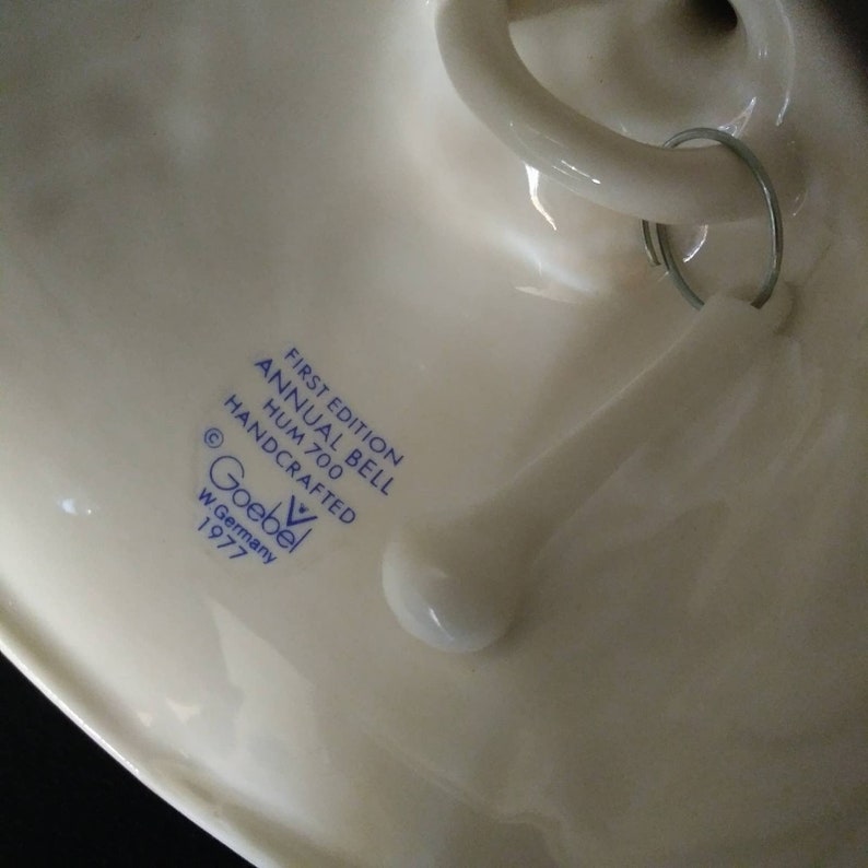 May include: A white porcelain bell with blue text that reads "First Edition Annual Bell Hum 700 Handcrafted &copy; Goebel W. Germany 1977".