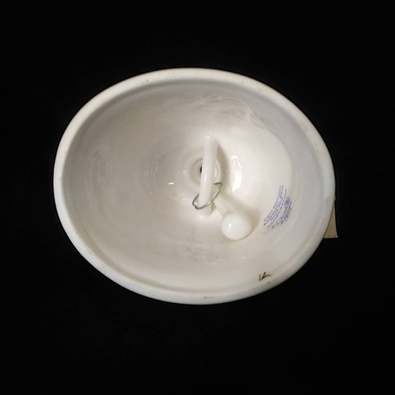 May include: White ceramic bell with a blue and white design on the bottom. The bell has a metal loop and a white knob for hanging.
