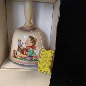 May include: A ceramic bell with a cream-coloured base and a colourful illustration of a boy playing an accordion with a bird perched on his shoulder. The bell is in a box with a yellow tag that reads "Welcome to the wonderful world of Goebel Collectors."