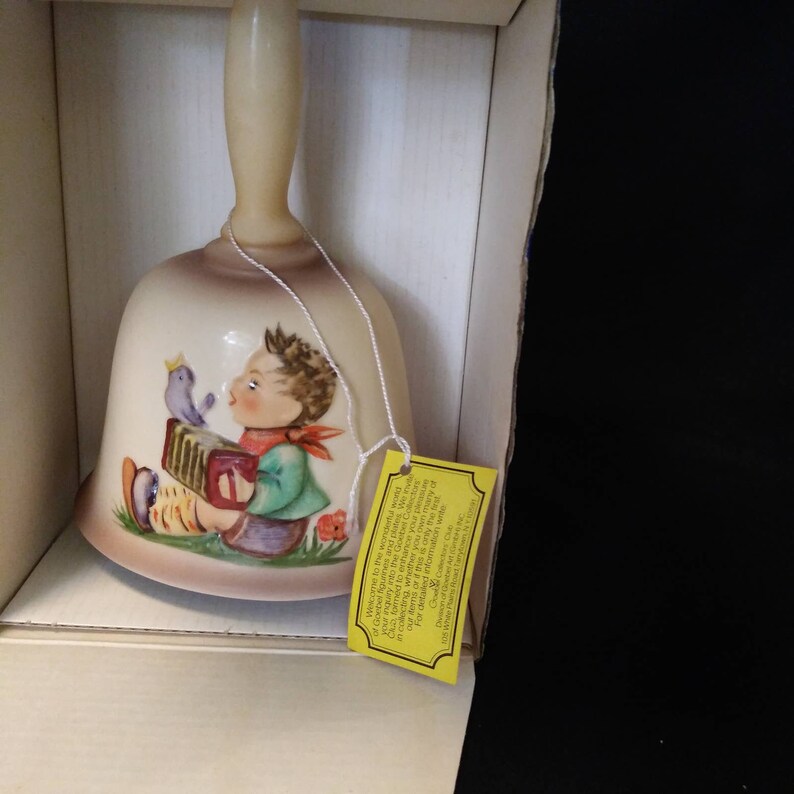 May include: A ceramic bell with a cream-coloured base and a colourful illustration of a boy playing an accordion with a bird perched on his shoulder. The bell is in a box with a yellow tag that reads "Welcome to the wonderful world of Goebel Collectors."