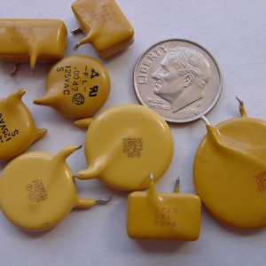 May include: A collection of eight yellow ceramic capacitors with various markings and sizes. The capacitors are arranged on a white surface. The largest capacitor is about the size of a US dime.