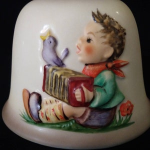 May include: A ceramic figurine of a boy playing an accordion with a purple bird perched on his shoulder. The boy is wearing a green shirt, red scarf, and blue trousers. The figurine is painted in a whimsical style with bright colours.