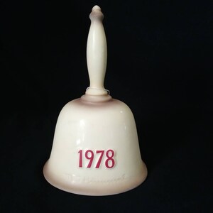 May include: A white ceramic bell with a brown glaze and a wooden handle. The bell has the year "1978" printed in red on the front.