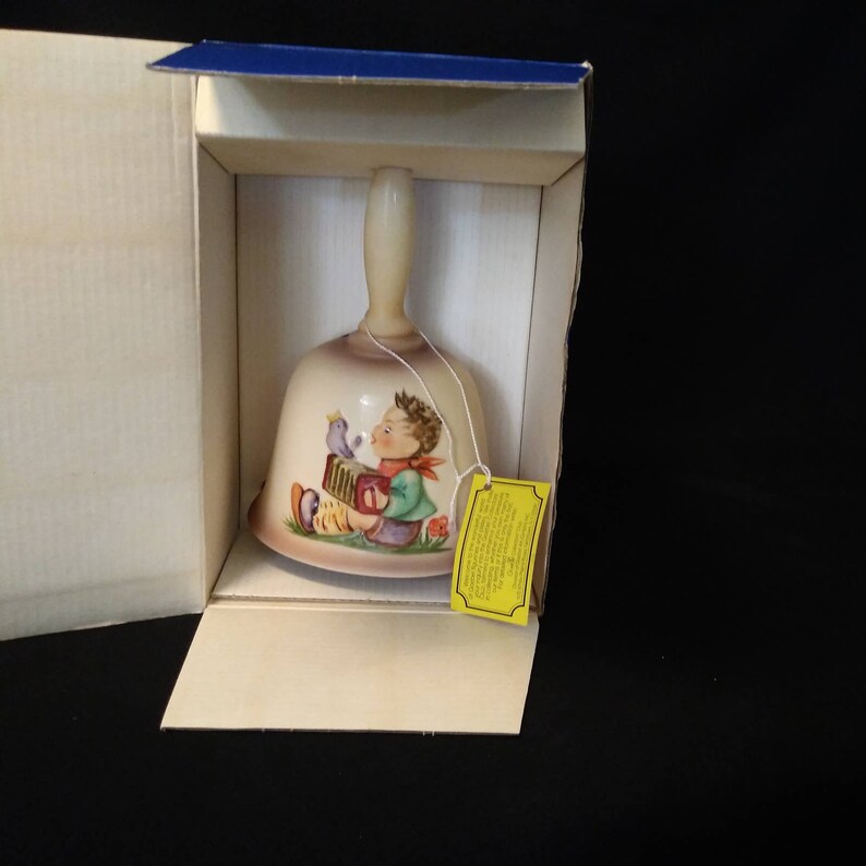 May include: A porcelain bell featuring a hand-painted design of a boy playing an accordion with a bird perched on his shoulder. The bell is in its original box with a yellow tag.