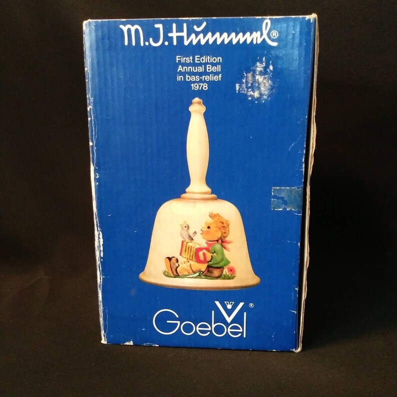 May include: A blue box containing a white bell. The bell has a white handle and a picture of a child playing an accordion with a bird on top. The box reads "M.J. Hummel First Edition Annual Bell in bas-relief 1978" and "Goebel".