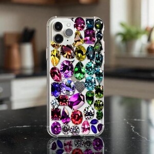 May include: A clear smartphone case decorated with a dense arrangement of multi-colored, faceted faux gemstones in various shapes including pear, round, oval, and rectangular cuts in shades of pink, purple, blue, green, yellow, and clear.