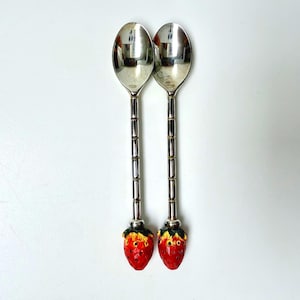 May include: Two silver-toned metal spoons featuring long, segmented handles and decorative strawberry-shaped finials at the base. The strawberries are painted in vibrant red with yellow and green accents at the top, contrasting with the polished metal.