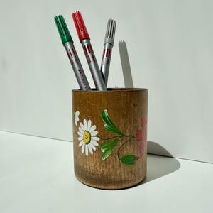 May include: A cylindrical wooden pencil holder featuring a hand-painted design of white daisies and green leaves. Three Faber-Castell markers in green, red, and grey are placed inside the container, which has a natural wood grain finish.