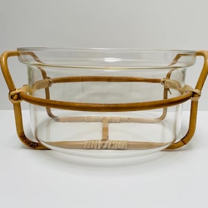 Puede incluir: Un cuenco de vidrio transparente con forma redondeada, sostenido por un marco de ratán natural. El marco presenta asas curvas y detalles tejidos, creando una pieza decorativa y funcional.