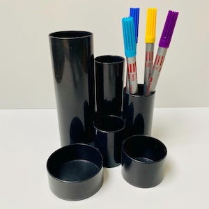 May include: Black plastic desk organiser with four round containers. The organiser holds three colourful markers with red and white stripes.