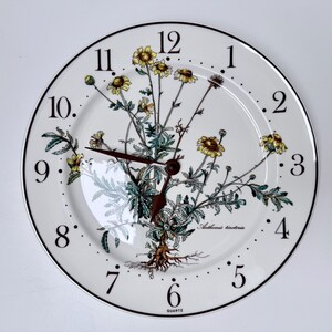 May include: A round wall clock with a white ceramic face featuring a botanical illustration of yellow Anthemis tinctoria flowers. The clock has black Arabic numerals from 1 to 12, dark brown hands, and the word QUARTZ printed at the bottom.