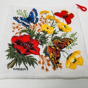 Op de afbeelding: Een witte katoenen ovenwant met een bloemenpatroon met rode, gele en blauwe vlinders. De ovenwant heeft een rode lusvormige handgreep en de tekst "KREIER" staat onderaan gedrukt.