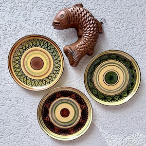 May include: A brown wooden fish-shaped mould with detailed scales and three round ceramic plates. The plates feature concentric circles and geometric patterns in shades of green, brown, and cream, arranged in a cluster around the fish mould.