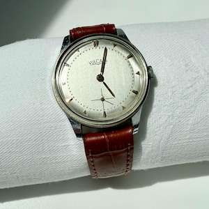 May include: A silver-toned wristwatch with a round face, featuring a brushed silver dial, gold-colored hour markers, and the text VULCAIN printed in black. The watch includes a brown leather strap with a textured, embossed crocodile-skin pattern.