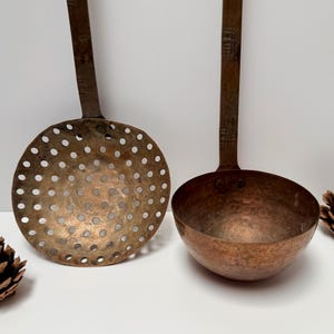 Puede incluir: Dos utensilios de cocina antiguos de cobre: un espumadera y un cucharón. La espumadera tiene una cabeza redonda y perforada. El cucharón tiene un cuenco profundo y redondeado. Dos piñas se colocan a ambos lados de los utensilios.
