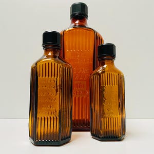以下が含まれることがあります： 黒いキャップが付いた琥珀色のガラス瓶が3本。瓶はリブ付きで、瓶には文字が書かれています。一番大きな瓶の文字は「LE REMEDE DE LA PASSE NON SERIEUX」。真ん中の瓶の文字は「JUST LE HALICEN HE HAS RUBY ALL DAY NON OTHER」。一番小さな瓶の文字は「INSUFFISAN NE PAS AVALER NON INGERIBLE」。