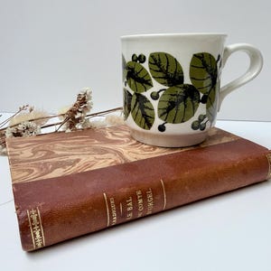 May include: A white ceramic mug featuring a repeating pattern of dark green leaves and small berries, resting on a brown leather-bound book titled Le Bal du Comte d'Orgel by Radiguet, accompanied by a sprig of dried white flowers.