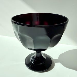 Op de afbeelding: Een zwarte glazen dessertkom met een steel en een ronde voet. De kom heeft een facetontwerp en een dieprood interieur. De kom staat op een wit oppervlak.