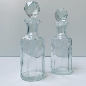 Puede incluir: Dos botellas de vidrio transparente con tapones geométricos. Las botellas están vacías y tienen una forma cuadrada con bordes redondeados.