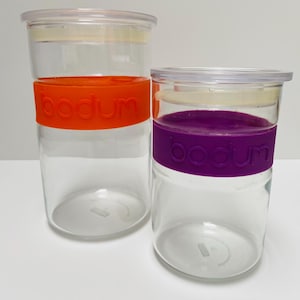 Op de afbeelding: Twee doorzichtige glazen opbergcontainers met transparante deksels. Eén heeft een oranje band met het woord "bodum" en de andere een paarse band met hetzelfde woord. De containers zijn cilindrisch en staan op een wit oppervlak.