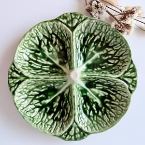 May include: A green ceramic serving dish with a four-sectioned design, resembling a cabbage leaf. The dish has a glossy finish and a central, raised, white circular area. The leaf-like sections are detailed with darker green veining.