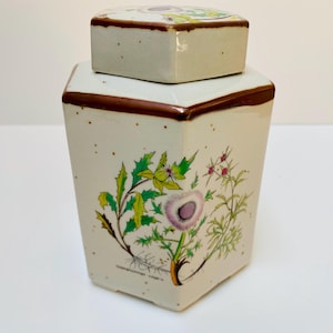Könnte beinhalten: Ein sechseckiger Keramiktopf mit passendem Deckel. Der Topf ist cremefarben mit braunen Zierleisten und einem Blumendesign mit grünen Blättern und lila Blüten. Der Deckel hat ein Blumendesign.