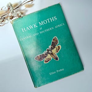 May include: A green hardcover book titled HAWK MOTHS of CENTRAL AND SOUTHERN AFRICA by Elliot Pinhey. The cover features a detailed illustration of a moth with brown, tan, and yellow patterned wings. Dried white flowers are visible at the top.