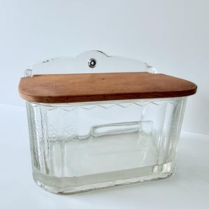 May include: Clear glass wall-mounted container with a wooden lid. The container has a rectangular shape with a textured surface. The lid is a warm brown colour. A small metal piece is attached to the top for hanging.