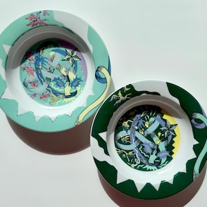 Può includere: Due ciotole decorative in ceramica. Una ciotola ha un bordo turchese con accenti bianchi e un design interno colorato. L'altra ciotola ha un bordo verde scuro con accenti bianchi e un design interno colorato. Entrambe le ciotole hanno un design artistico unico.