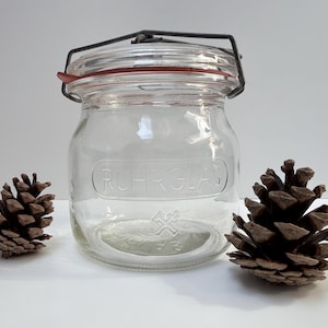 Op de afbeelding: Een heldere glazen pot met een metalen scharnierend deksel en een rode rubberen afdichting. De pot heeft het woord "RUHRGLAS" in reliëf op de voorkant. Twee bruine dennenappels staan aan weerszijden.