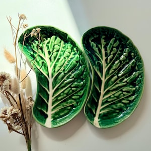 May include: Two green ceramic leaf-shaped plates with white veins, arranged side by side. The plates have a glossy finish and are set against a white background. Dried flowers are placed to the left of the plates.