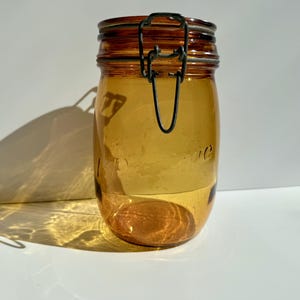 Könnte beinhalten: Bernsteinfarbenes Glasgefäß mit Metallverschluss. Das Gefäß hat eine abgerundete Form und einen klappbaren Metalldeckel. Das Gefäß ist leer und steht auf einer weißen Oberfläche.