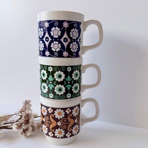 May include: Three stacked ceramic mugs with white handles. The top mug is navy blue with a white and pink floral pattern. The middle mug is green with white and teal floral designs. The bottom mug is brown with white daisies and orange accents.