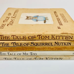 以下が含まれることがあります： ベージュ色の表紙の、ヴィンテージの子供向けの本の束。「The Tale of Tom Kitten」、「The Tale of Squirrel Nutkin」、「The Tale of Mr. Tod」などのタイトル。一番上の本には、青い服を着た猫のイラストが描かれています。