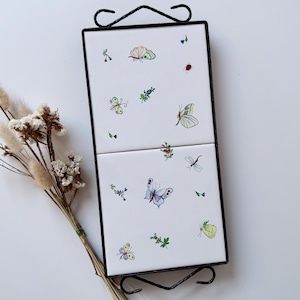May include: A black metal wall hanging with two white ceramic tiles decorated with colourful watercolour illustrations of butterflies, flowers, and insects. A dried flower arrangement is placed to the left of the wall hanging.