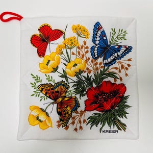 Op de afbeelding: Een witte katoenen ovenwant met een bloemenpatroon met rode, blauwe en gele vlinders, gele en rode bloemen en groene bladeren. De tekst "KREIER" is onderaan in het zwart gedrukt.