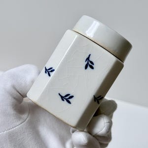 May include: A rectangular ceramic canister with a glossy off-white crackle glaze finish. The canister features a repeating pattern of small, dark blue leaf motifs scattered across its surface and includes a matching removable lid.