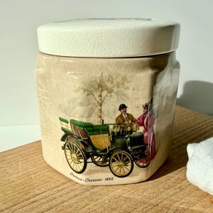 Può includere: Un barattolo in ceramica con coperchio, raffigurante una scena di auto d'epoca. Il barattolo ha uno sfondo beige con un'illustrazione dettagliata di un'auto classica, un albero e delle figure. Il barattolo è su una superficie di legno.