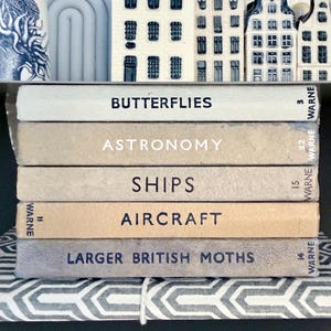 Puede incluir: Una pila de libros antiguos con títulos como "Butterflies", "Astronomy", "Ships", "Aircraft" y "Larger British Moths". Los libros son de varios tonos beige y gris. Casas decorativas de cerámica se encuentran encima de los libros en una estantería.