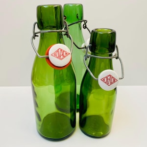 Könnte beinhalten: Drei grüne Glasflaschen mit Bügelverschlüssen. Die Flaschen haben ein weißes und rotes Logo auf der Vorderseite. Die Flaschen sind unterschiedlich groß und in einer Gruppe angeordnet.
