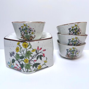 May include: A ceramic fondue pot with a square base and a matching bowl, accompanied by three additional small bowls. Each piece is off-white with brown speckles and features a colourful botanical illustration of yellow flowers and red berries.