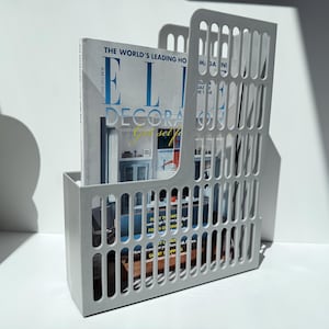 May include: A white metal magazine holder featuring a vertical slotted pattern on the sides and front. The holder contains a copy of Elle Decoration magazine with visible text including the title and various headlines on the cover.