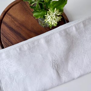 May include: White linen towel with an embroidered monogram and decorative border. The towel is folded and placed on a wooden tray with a small vase of flowers. The overall aesthetic is clean and elegant.