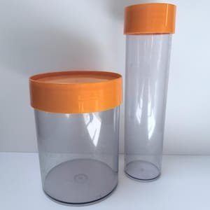 Könnte beinhalten: Zwei transparente Kunststoffbehälter mit orangefarbenen Deckeln. Einer ist zylindrisch und hoch, der andere kürzer und breiter. Beide Behälter sind leer und stehen auf einer weißen Oberfläche vor einem weißen Hintergrund. Sie sind wahrscheinlich für die Aufbewahrung oder Präsentation.