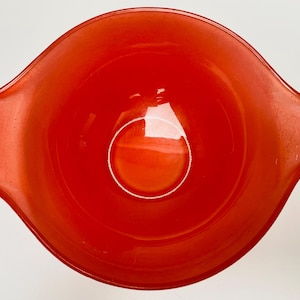 May include: A vibrant, red vintage mixing bowl with two handles. The bowl is made of glass and has a rounded shape. The interior of the bowl is a lighter shade of red, and the exterior is a deeper, more saturated red.
