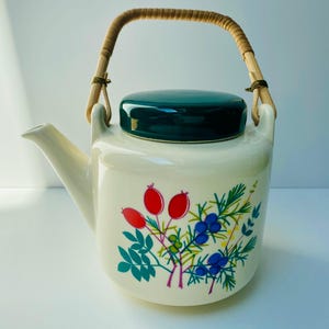 Può includere: Teiera in ceramica color crema con coperchio verde scuro e manico intrecciato. La teiera è decorata con un motivo floreale con bacche rosse, bacche blu e foglie verdi. Il beccuccio è sul lato sinistro.