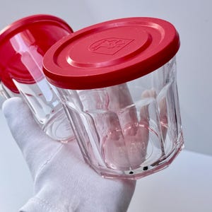 May include: Two clear glass jars with faceted sides and bright red plastic lids. Each lid features a circular embossed design with a central logo. The glass containers have a cylindrical shape with vertical ridges and a clear, transparent finish.