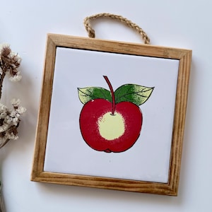 Puede incluir: Arte mural cuadrado con marco de madera que presenta una ilustración de una manzana roja con hojas verdes y un centro amarillo. La obra de arte tiene un marco de madera natural y una cuerda trenzada para colgar.