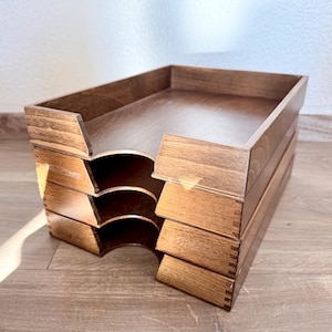 Könnte beinhalten: Ein Stapel von drei Schreibtisch-Organisatoren aus Holz in einem warmen braunen Farbton. Jedes Fach hat eine rechteckige Form mit abgerundeten Aussparungen für einen einfachen Zugriff. Die Holzmaserung ist sichtbar, und die Konstruktion weist Schwalbenschwanzverbindungen auf.