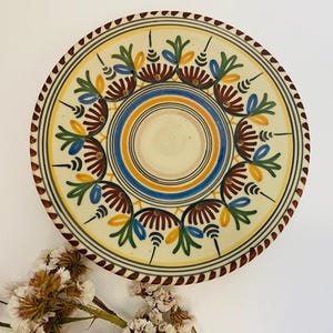 Puede incluir: Plato de cerámica decorativo con base crema y diseños florales pintados a mano en tonos marrón, azul, verde y amarillo. El plato presenta un borde detallado y un diseño central, perfecto para exhibir o servir.